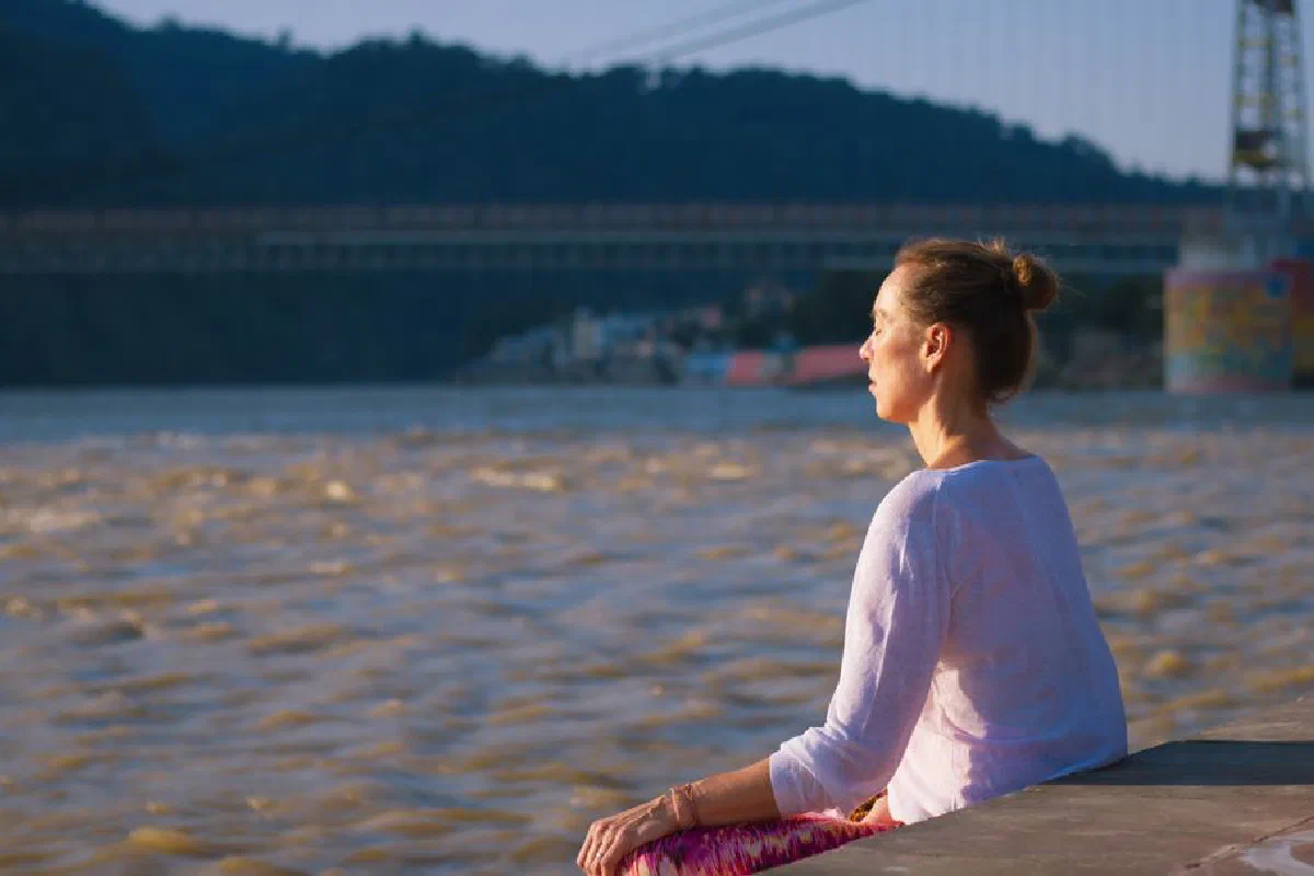 Woman meditating near Ganges River in Rishikesh, India with a bridge in the background.
