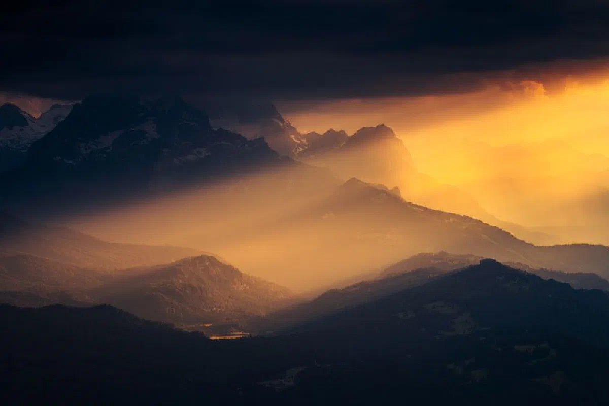 Sunset casting golden rays over majestic mountain peaks with dramatic clouds.