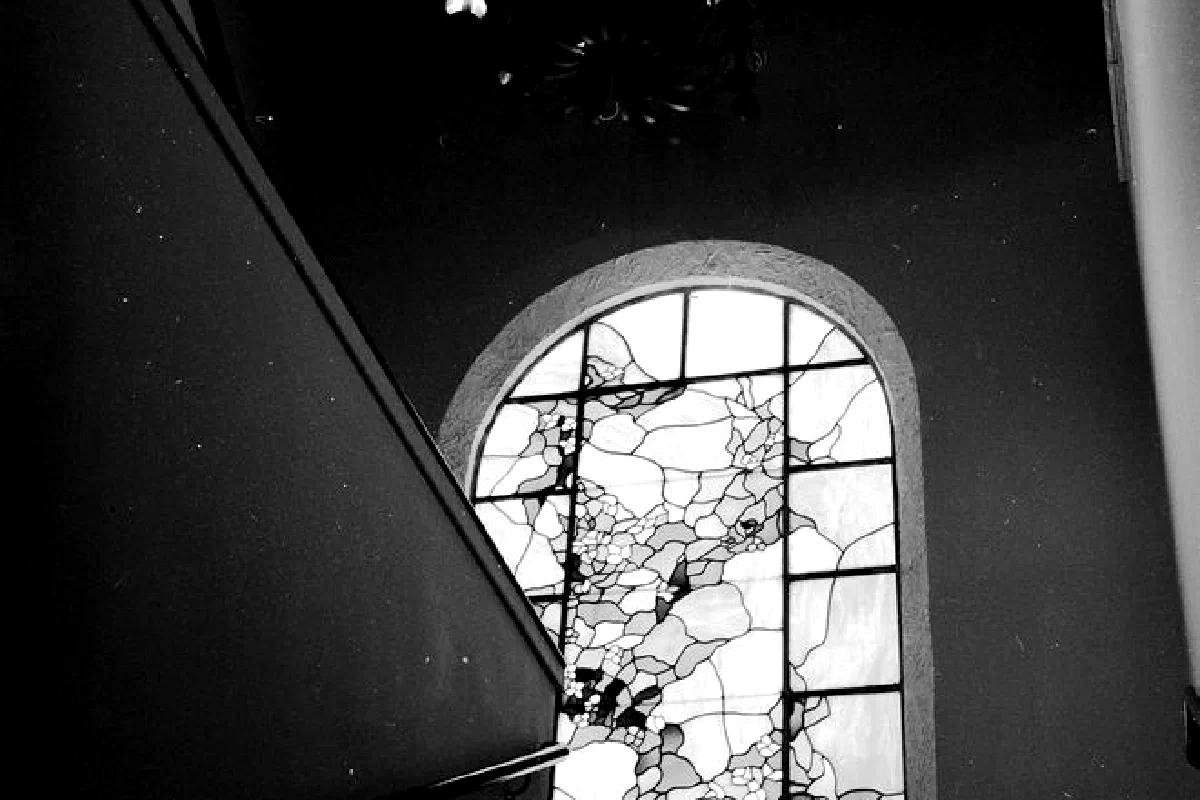 Monochromatic image of an arched stained glass window with chandelier above.