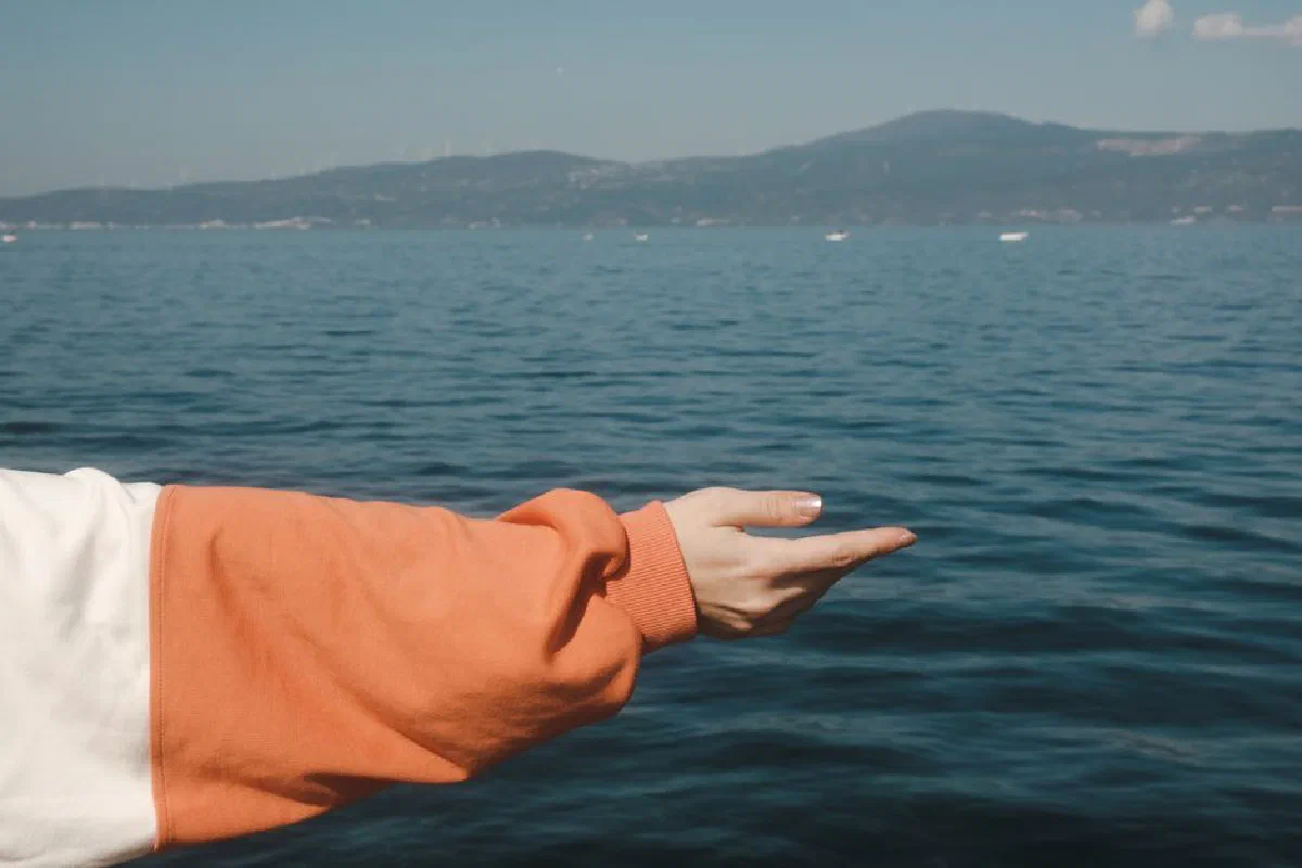 A person's hand reaching out over a tranquil lake with mountains in view, offering a sense of calm and contemplation.