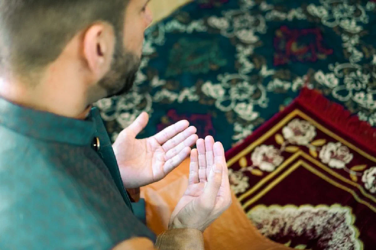 A man in traditional attire is praying on a beautifully decorated prayer mat indoors.