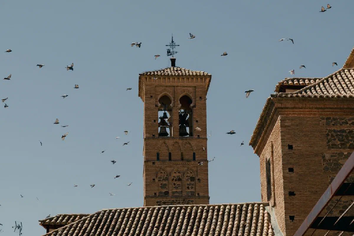 Birds fly around a historic bell tower in Toledo, Spain, showcasing Moorish architecture under a clear sky.