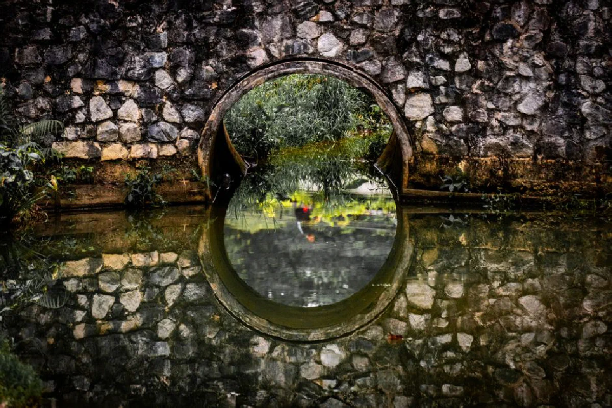 A picturesque stone arch bridge reflecting in a calm water surface, surrounded by greenery.