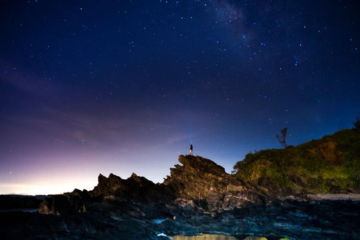 A lone person stands on rocky terrain beneath a stunning starry night sky by the ocean.