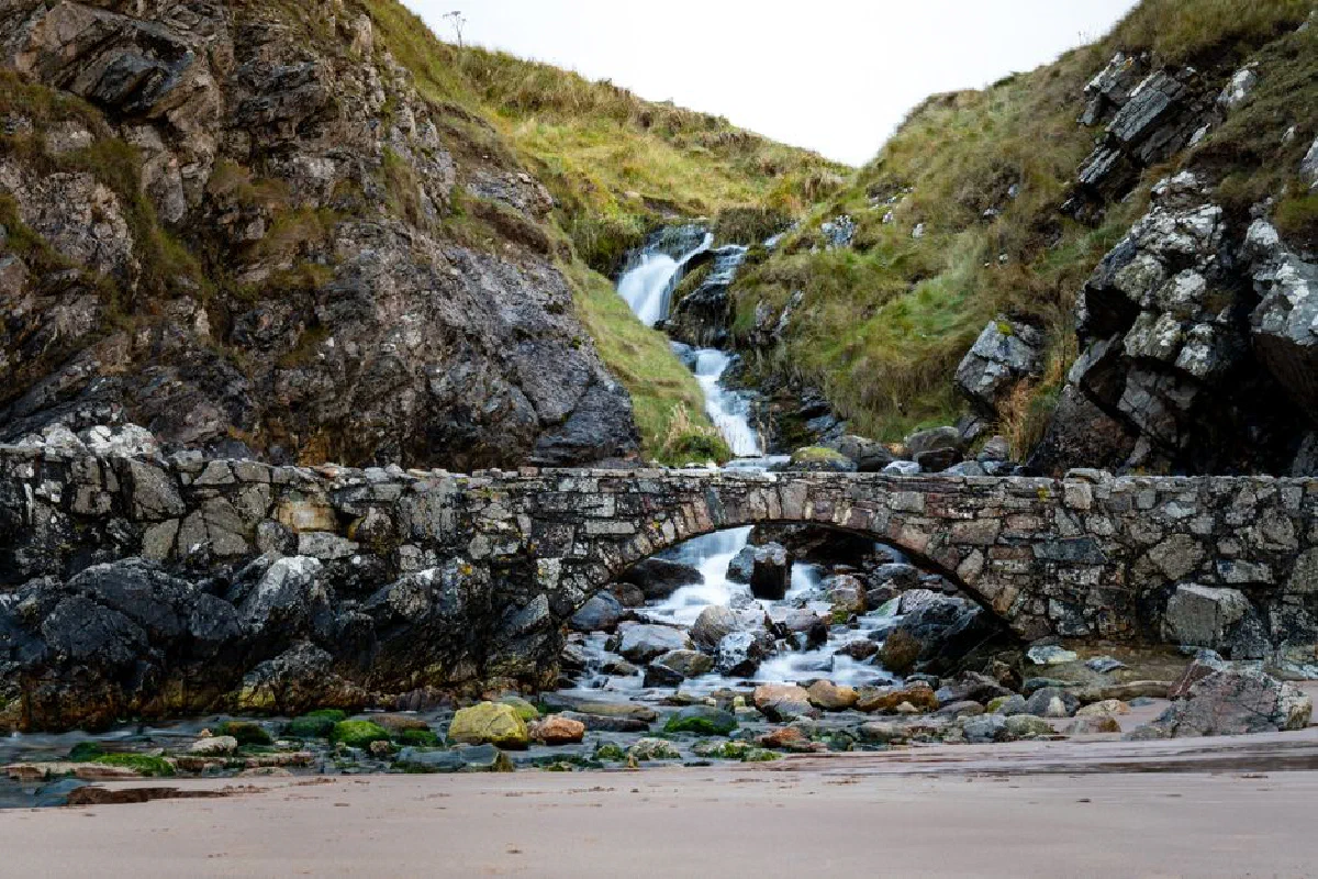 Stone bridge over a picturesque stream with a rocky hillside and waterfall in the background.