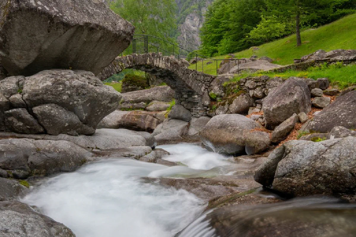 A serene scene of a stone bridge over a flowing creek surrounded by lush greenery in Cevio, Switzerland.