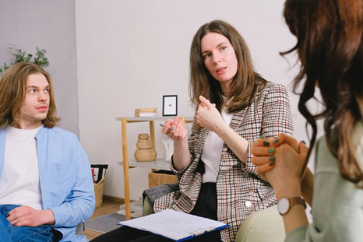 Thoughtful woman psychologist in stylish clothes siting with clipboard and talking with couple in light room