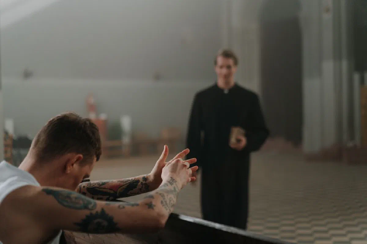A tattooed man prays in a church as a priest observes, capturing faith and reflection indoors.