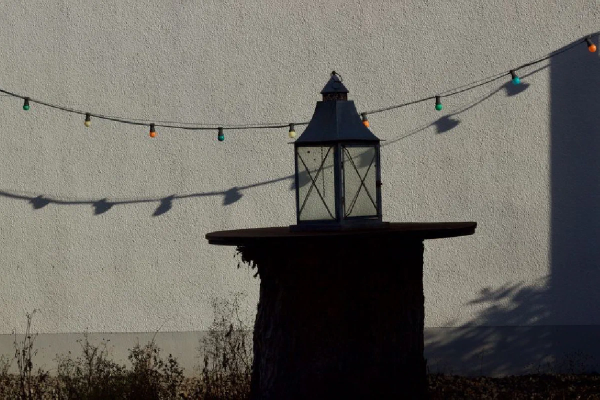 A rustic lantern on a tree stump with colorful string lights casting shadows against a white wall.