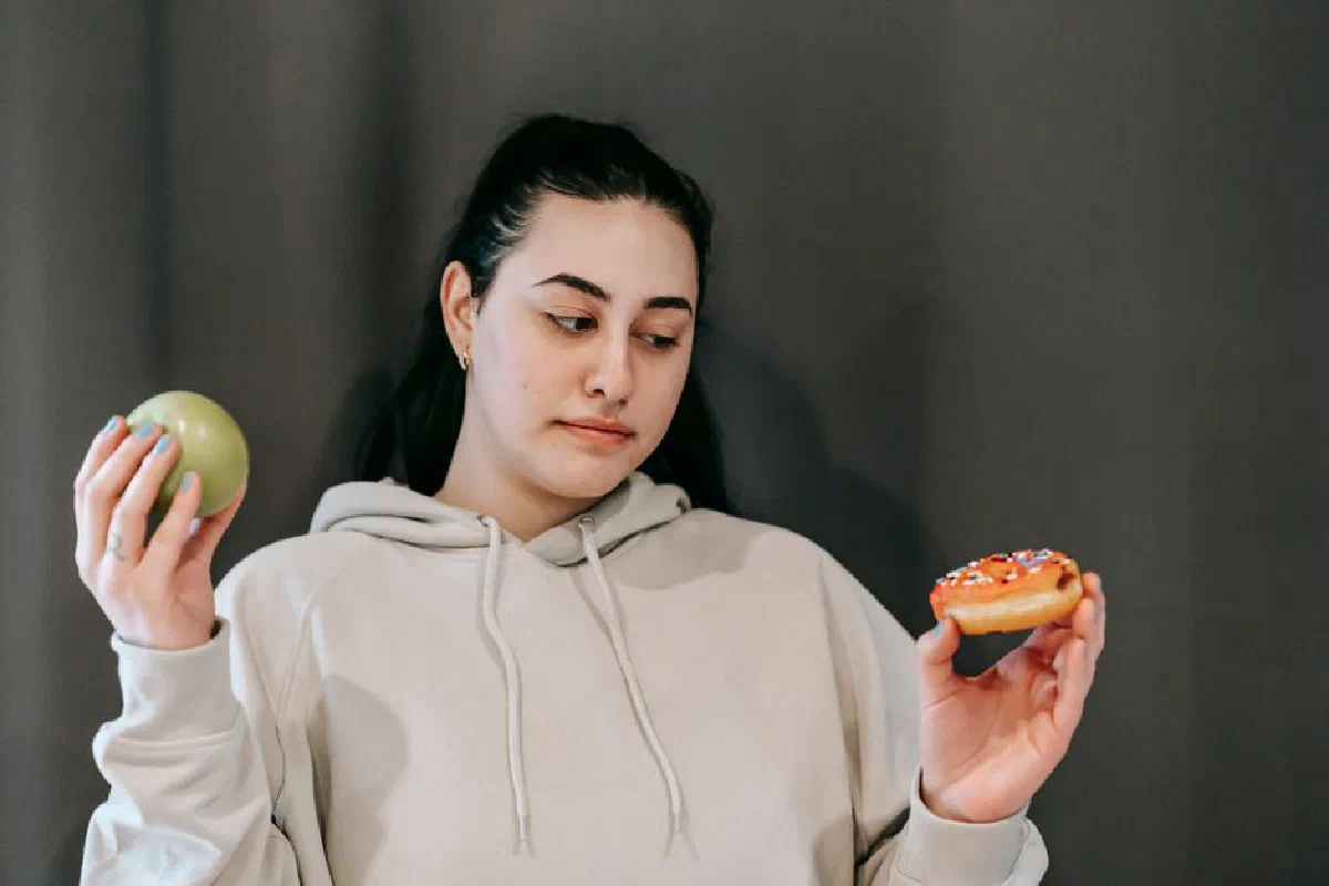 Contemplative plump female in hoodie choosing between ripe green apple and delicious donut in studio