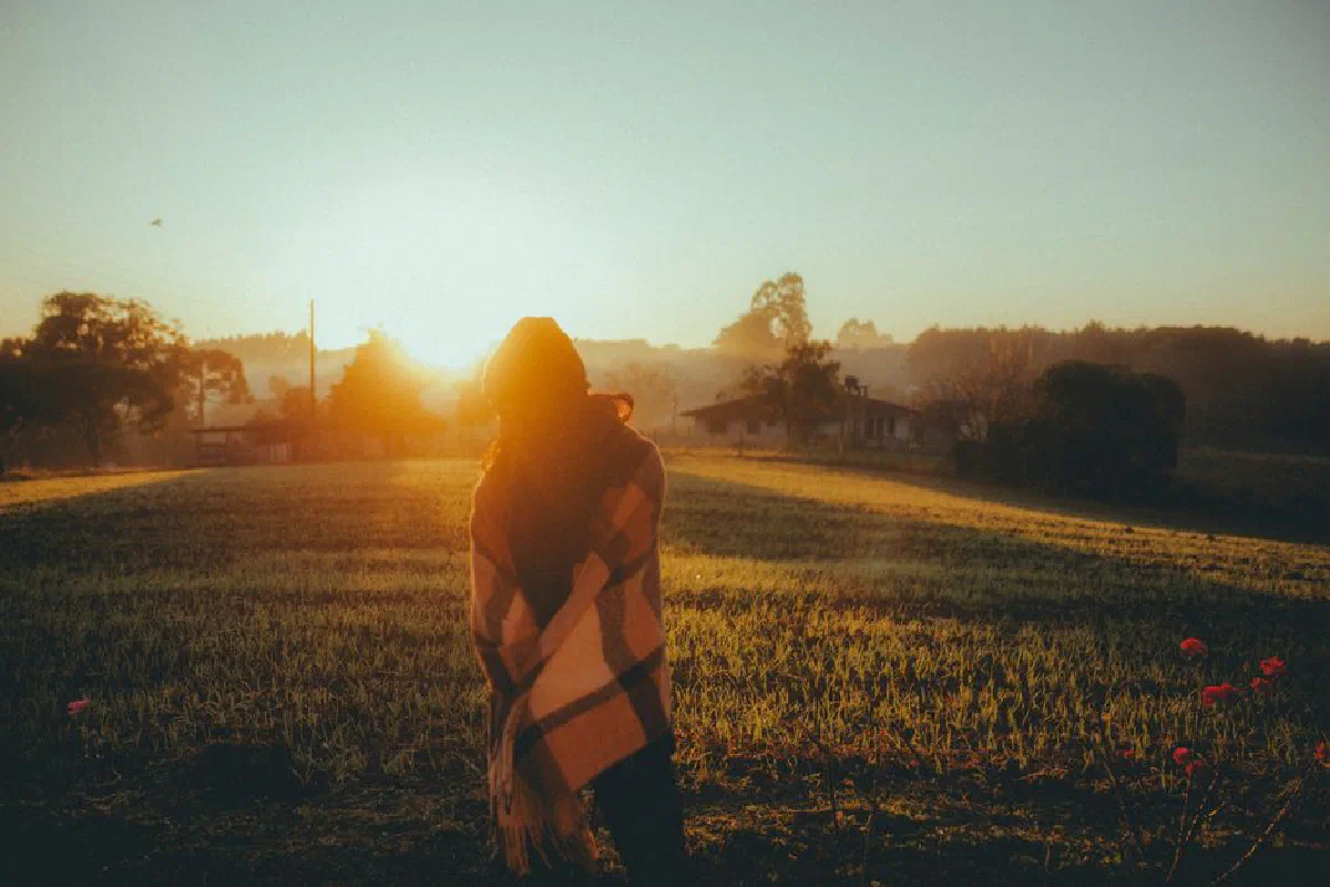 A woman wrapped in a blanket stands in a rural field with a beautiful sunset backdrop.