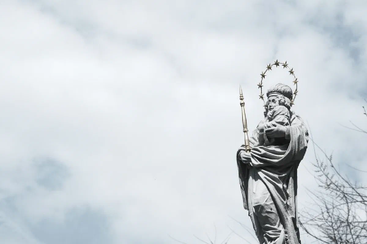 A striking statue of a saint with a halo and scepter set against a dramatic cloudy sky.
