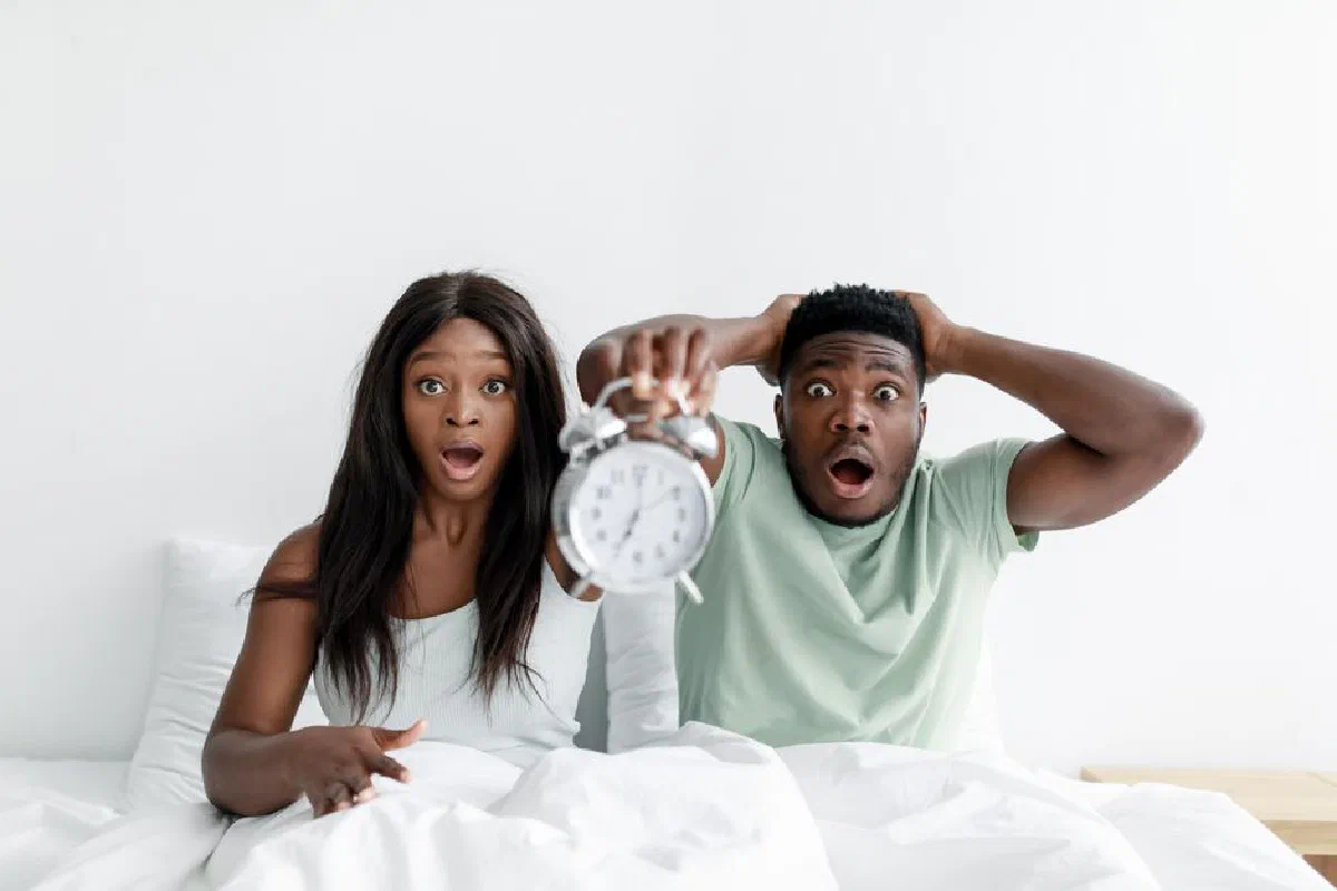 A surprised couple holding an alarm clock while sitting in bed, expressing shock.