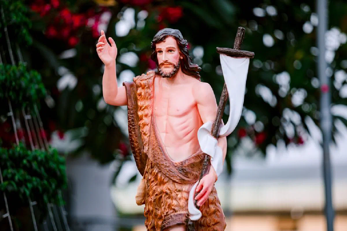 A detailed statue of John the Baptist holding a cross, set against a blurred outdoor backdrop.