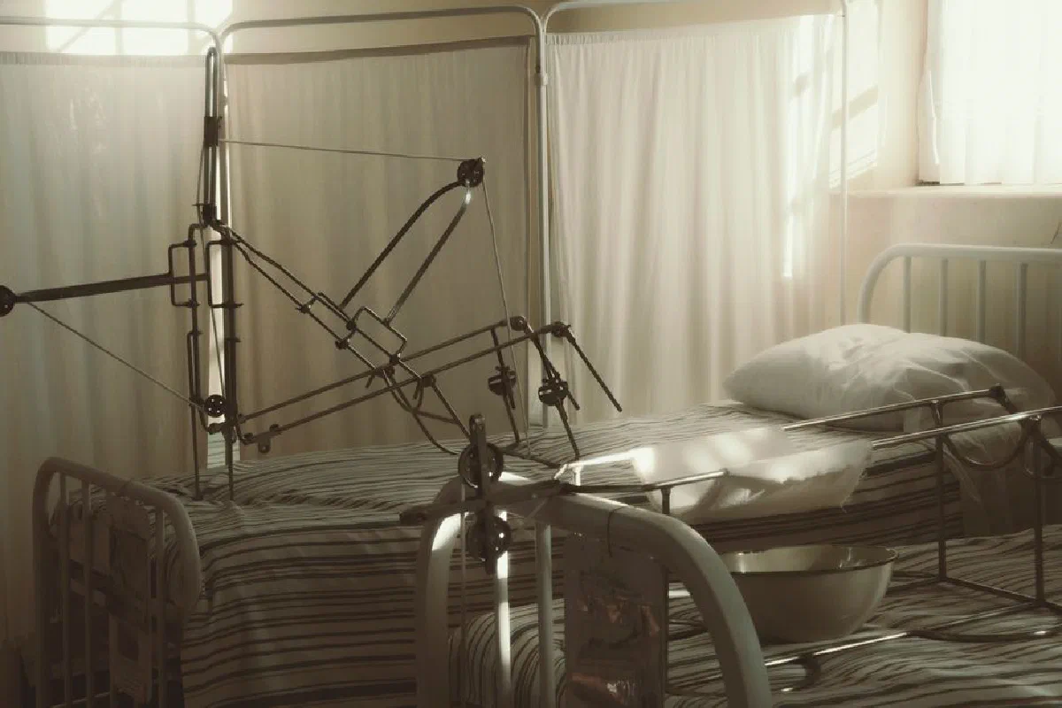 An empty hospital bed surrounded by medical equipment in a quiet, sunlit room.