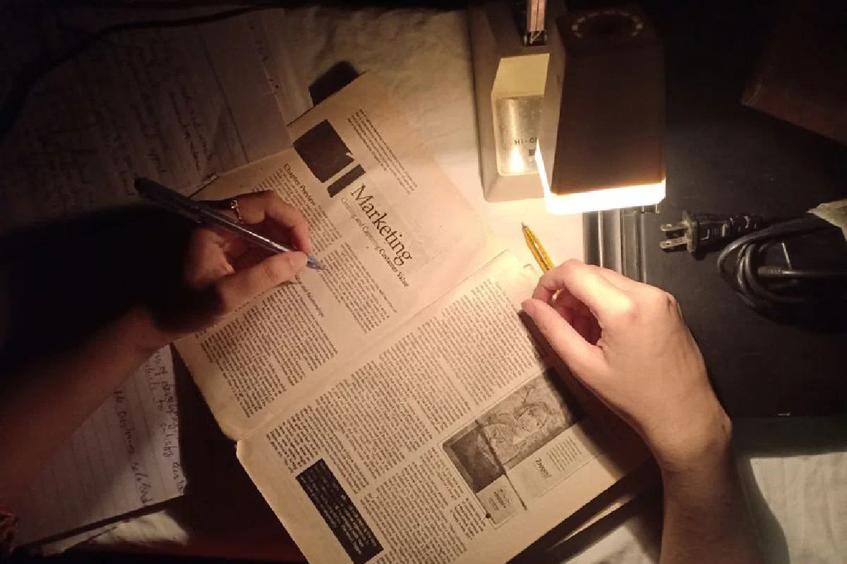 Hands writing on a marketing book under the warm glow of a desk lamp at night.