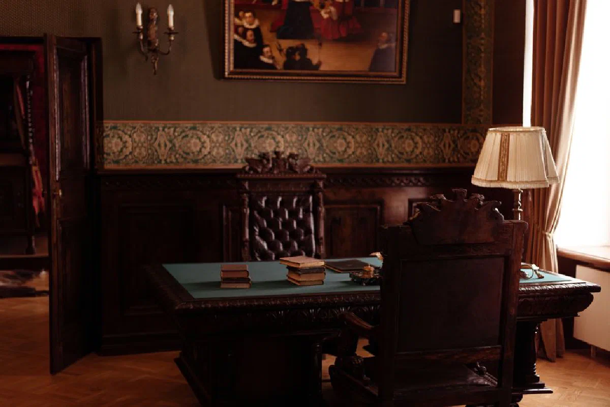 An ornate study room featuring antique wooden furniture, books, and a classic lamp.