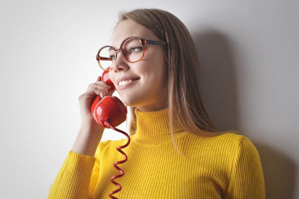 Woman in yellow sweater smiling while talking on a vintage red phone against a white wall.