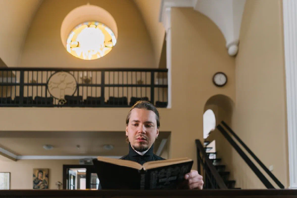 A priest reads a holy book inside a church under a stained-glass window.