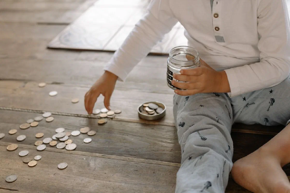 A young child collects coins into a jar on a wooden floor, symbolizing savings.