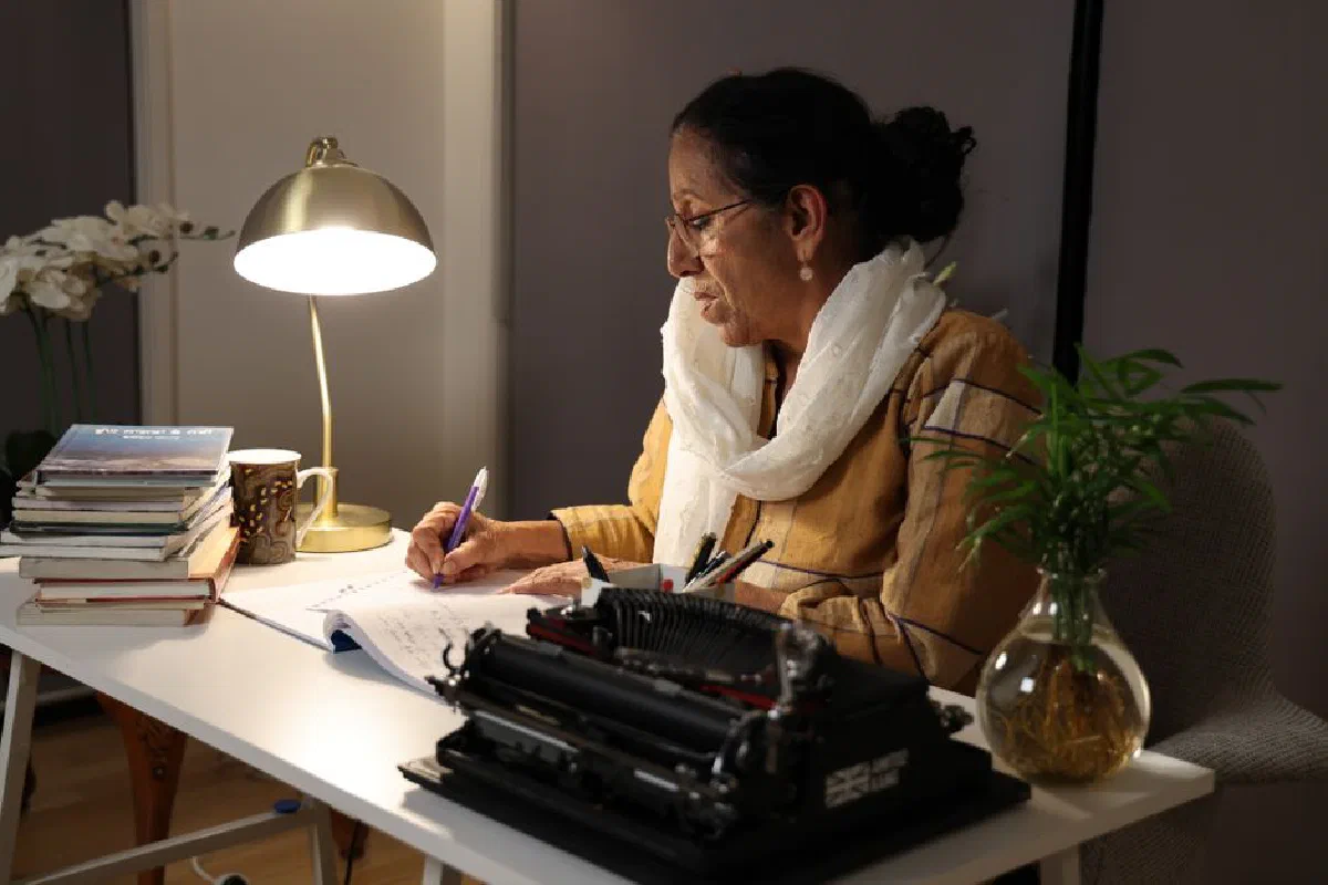 Senior woman writing at home desk with a typewriter and lamp.