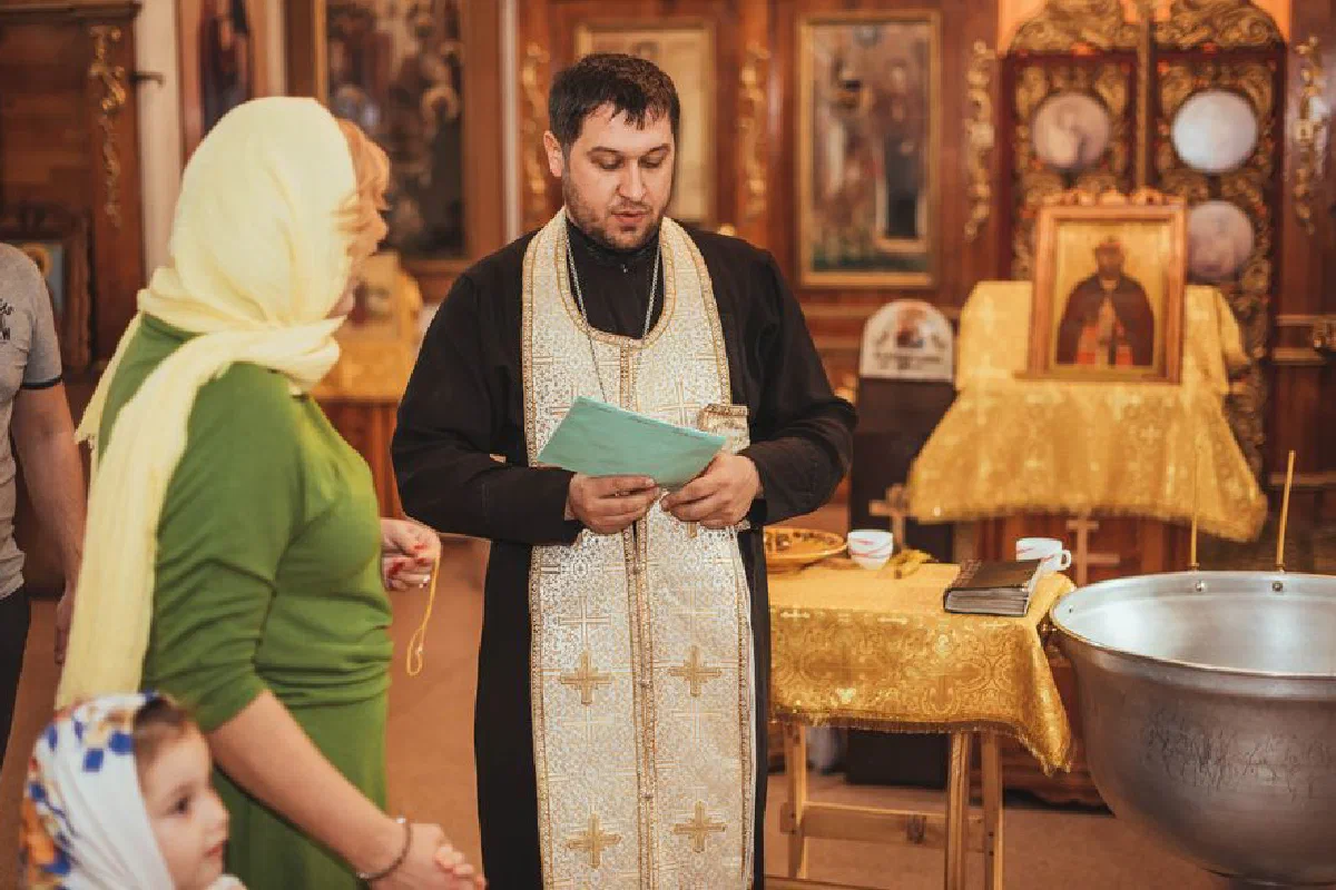 A priest conducting a baptism ceremony with family inside an Orthodox church.
