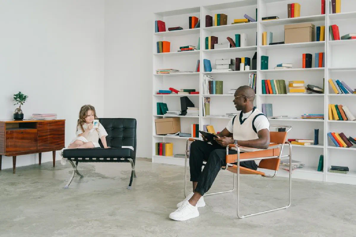 African American man teaching caucasian girl in a modern classroom with bookshelves.