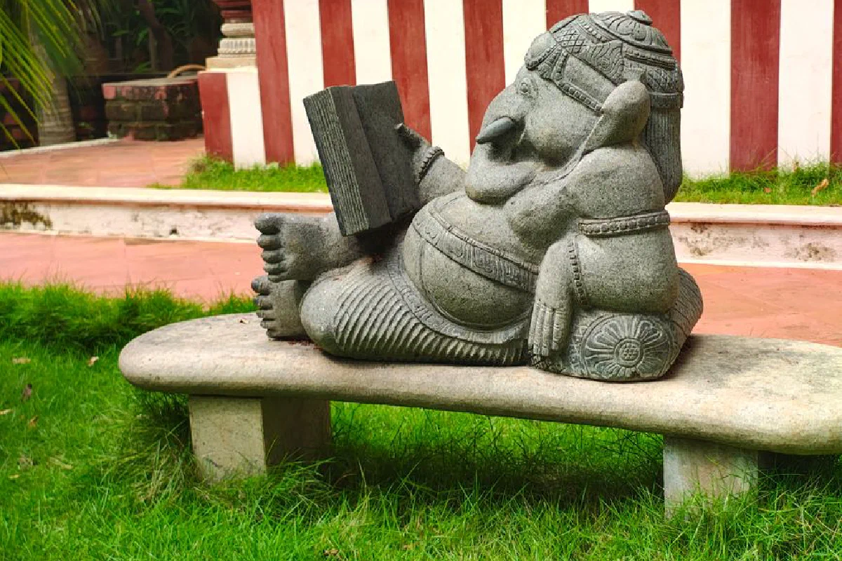 Stone statue of Ganesha relaxing and reading outdoors on a stone bench with lush grass.