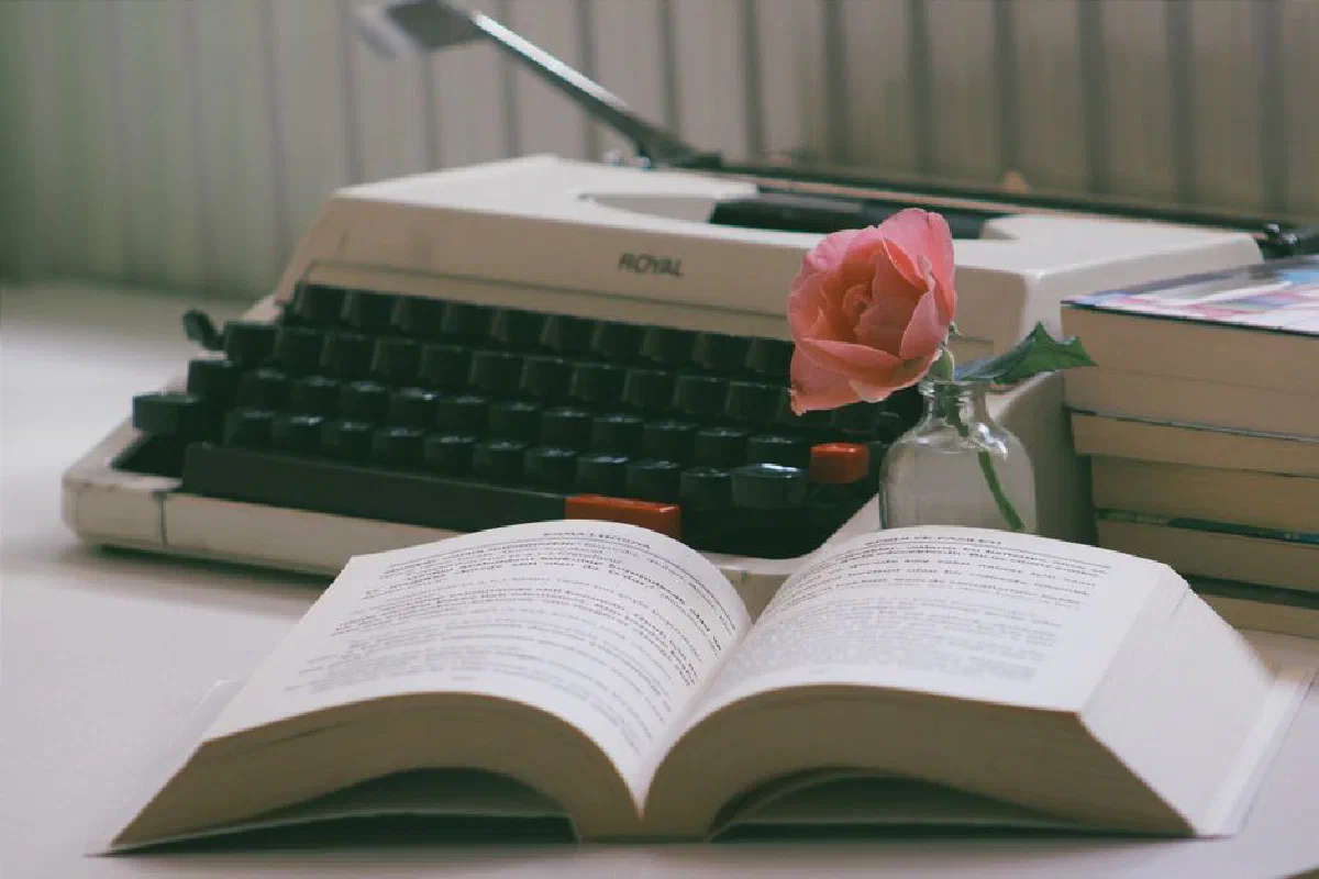 Cozy vintage setting with a typewriter, open book, and pink rose in a vase.