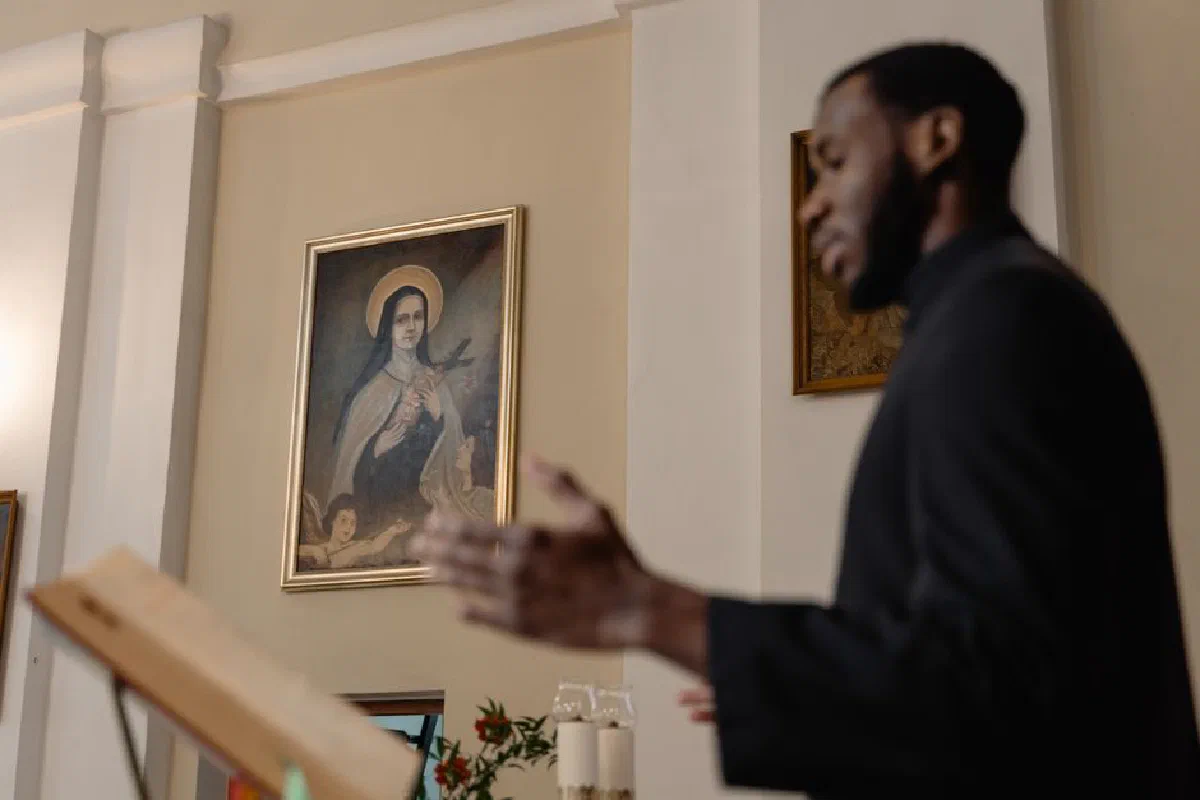 A priest delivers a sermon inside a church with religious art in the background.