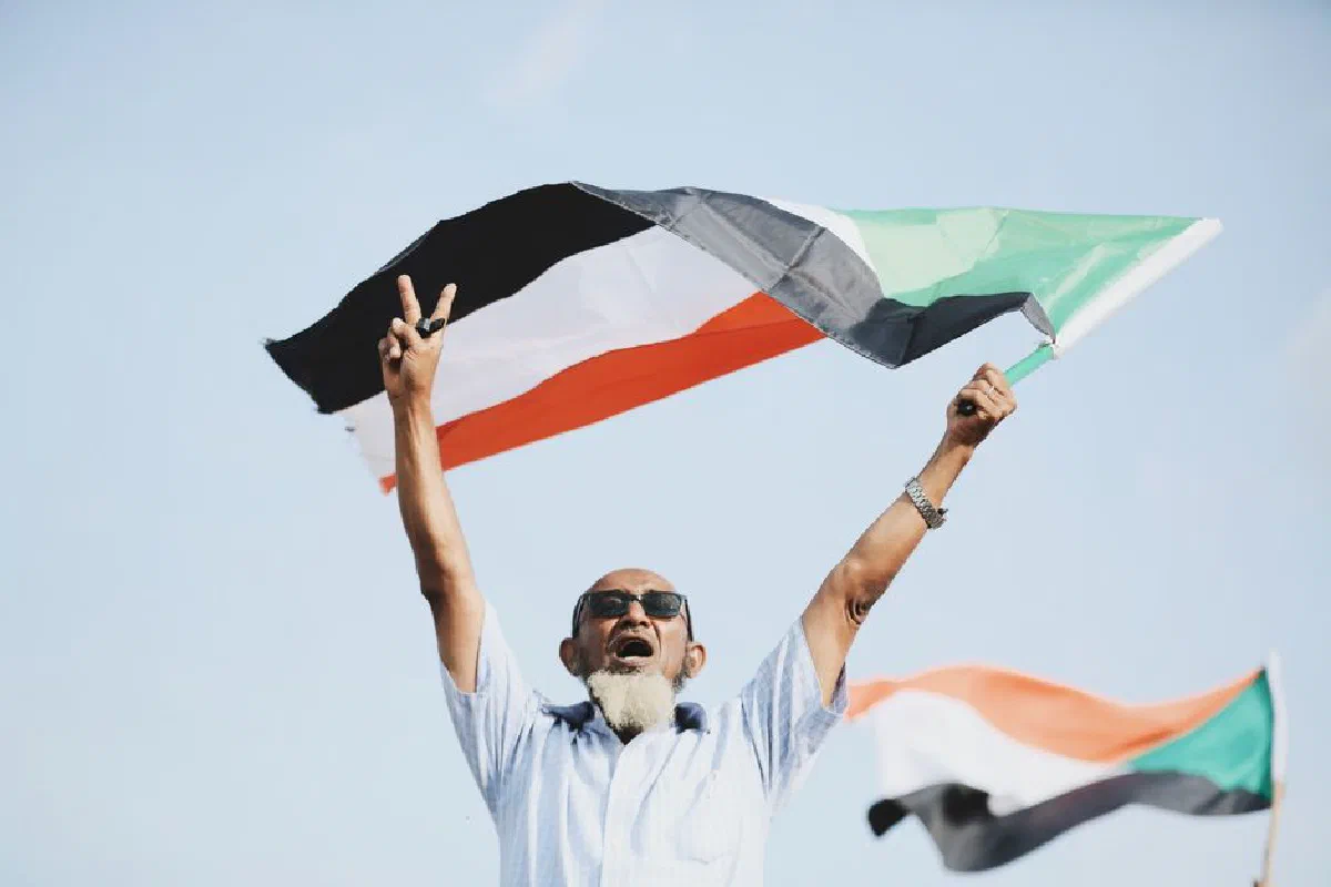 A joyful man holds the Sudanese flag, celebrating outdoors with a victory sign.
