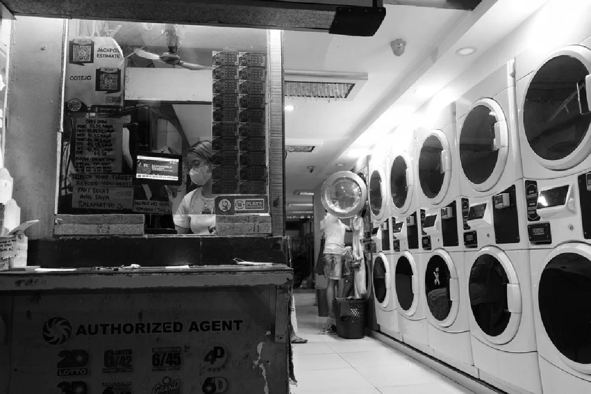 Black and white photo of a laundromat interior with a worker behind a counter. Captures city life.
