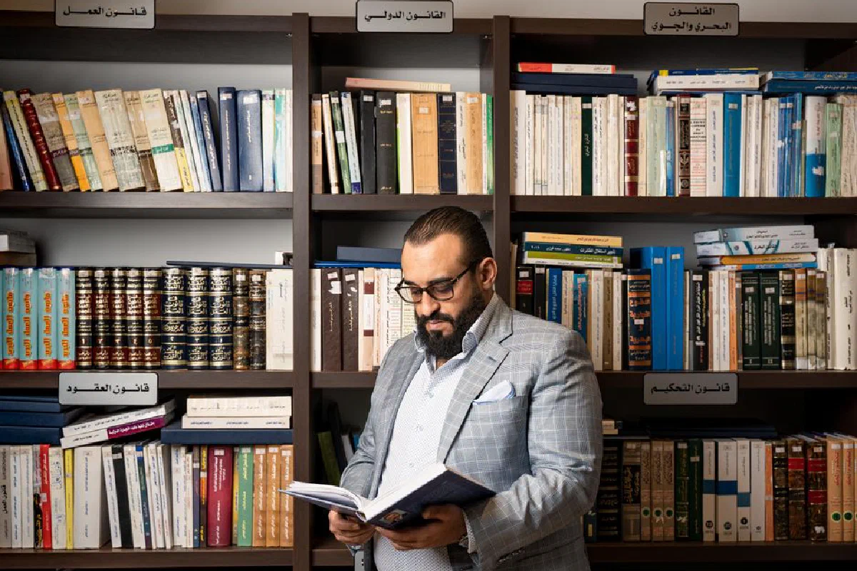 A bearded businessman in a suit reads a book in a library with shelves full of books.