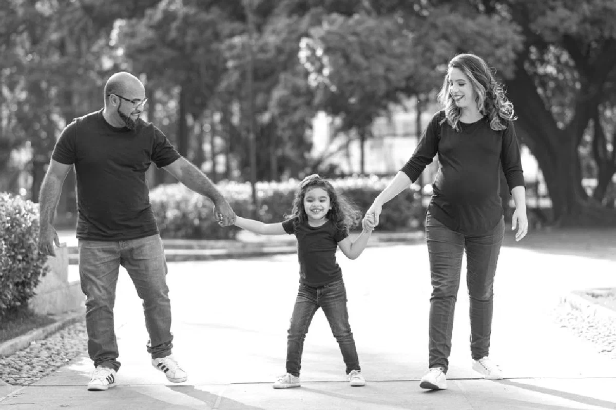 Happy family including a pregnant woman, man, and girl walking in a park holding hands.
