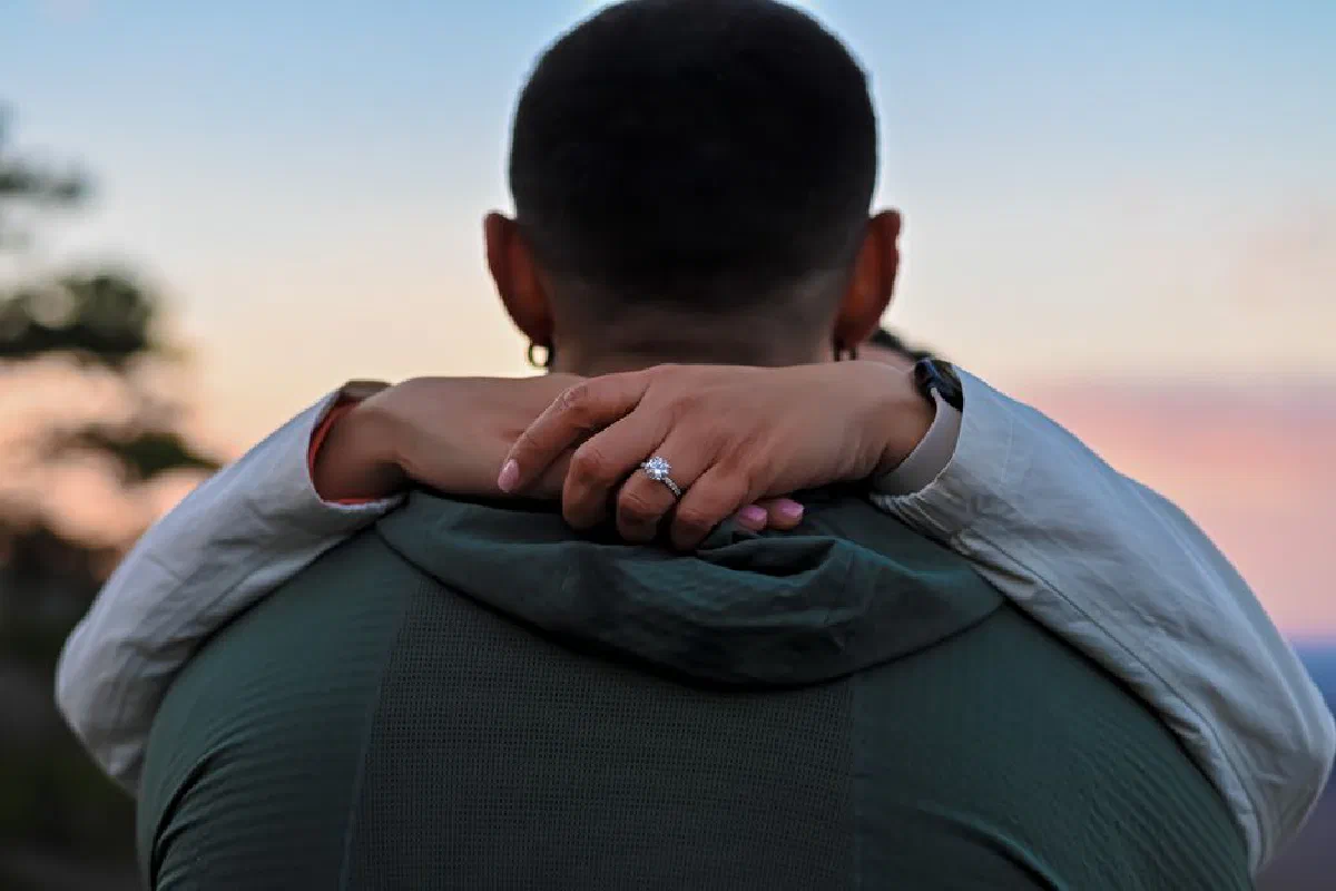 A couple embracing outdoors at twilight, with a focus on a woman's engagement ring.