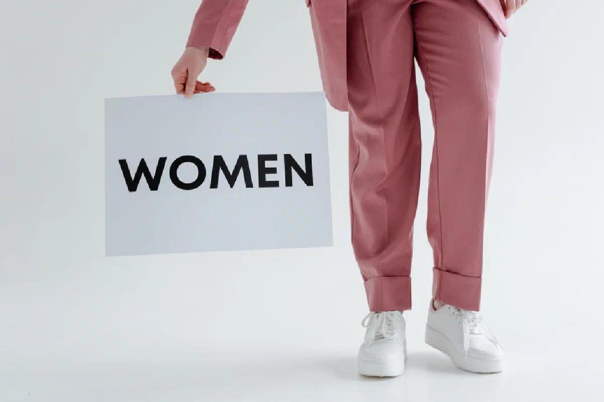 A woman in pink pants holds a 'WOMEN' sign in a minimalist studio with a white background.
