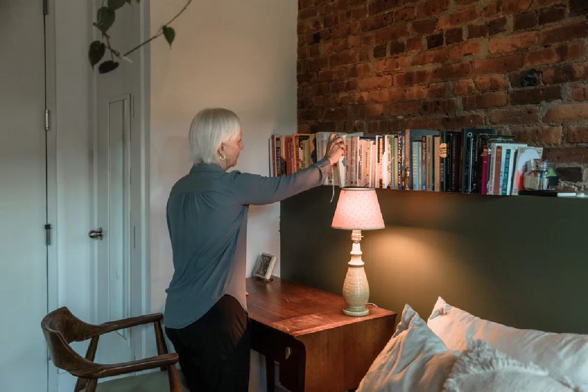 A senior woman arranging books on a shelf beside a desk lamp in a warm, cozy room.