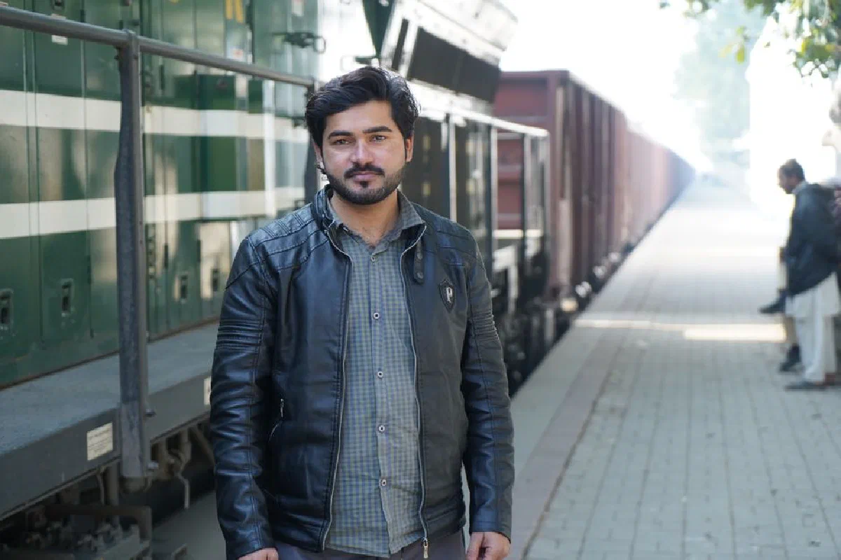A man in a black leather jacket poses confidently next to a train on a sunny day.
