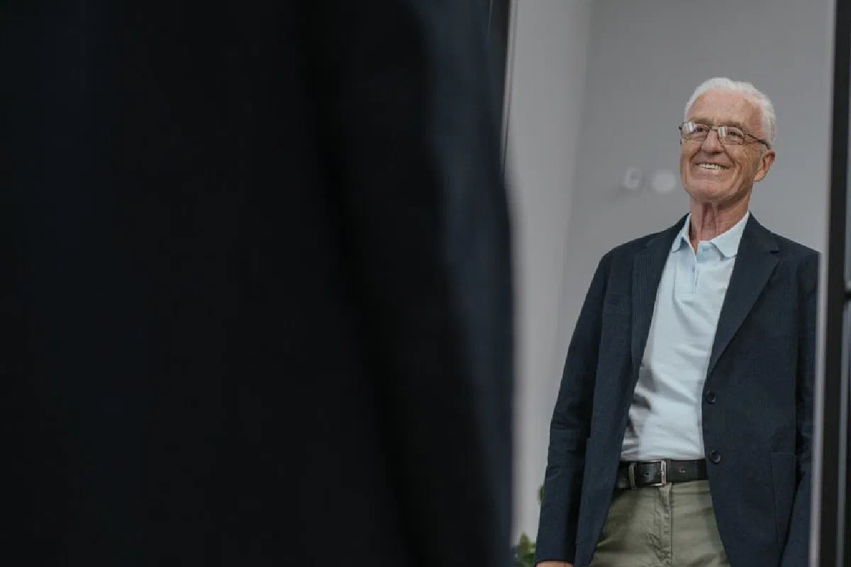 Senior man with eyeglasses smiling at his reflection while wearing a blue coat indoors.