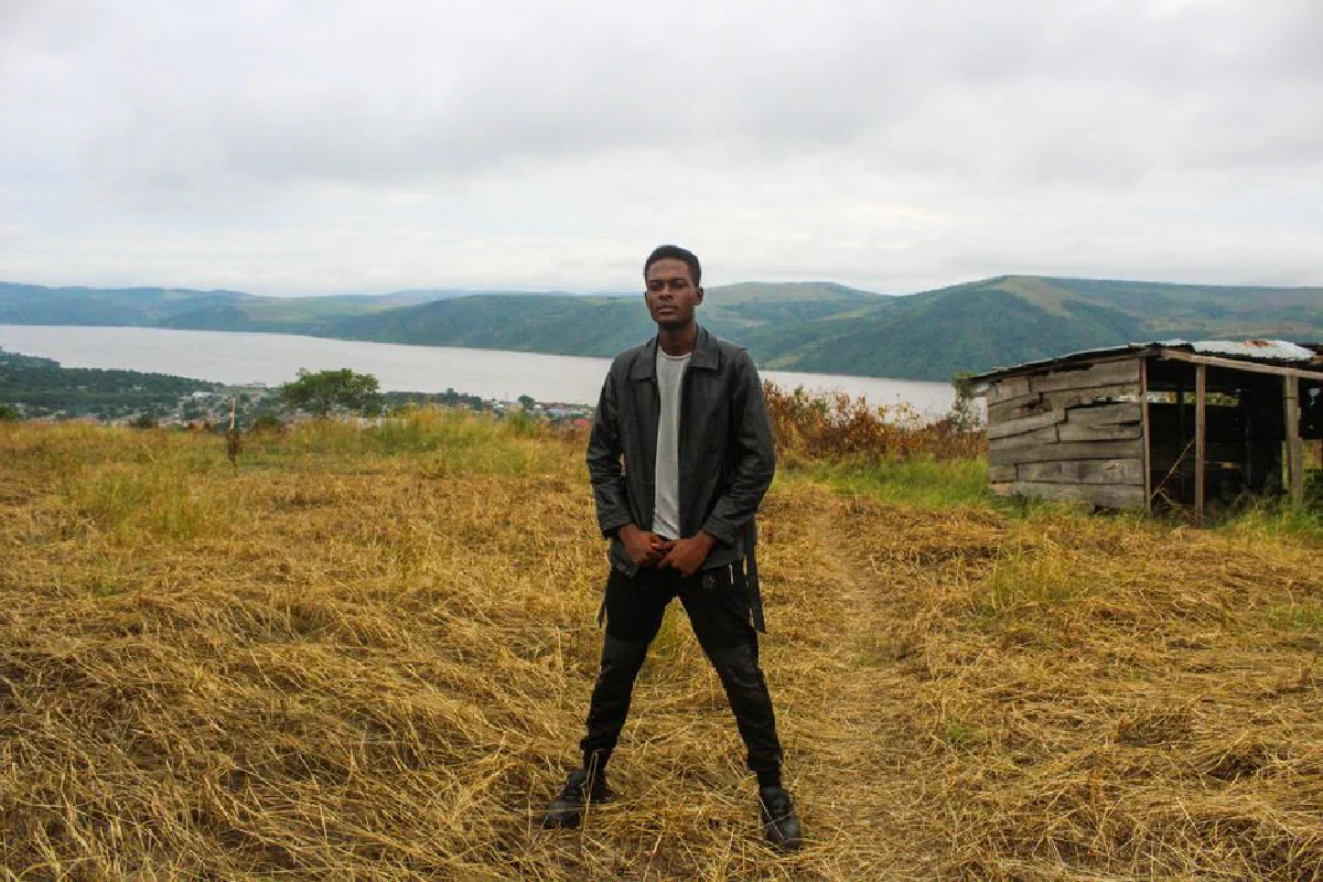 Young man in leather jacket posing outdoors near river and hills in Kinshasa.