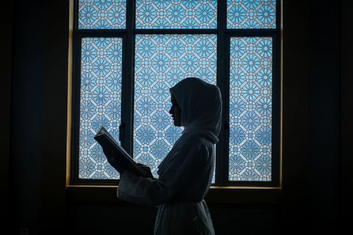 A Muslim woman reads the Quran by a decorative window, capturing a serene and spiritual moment.