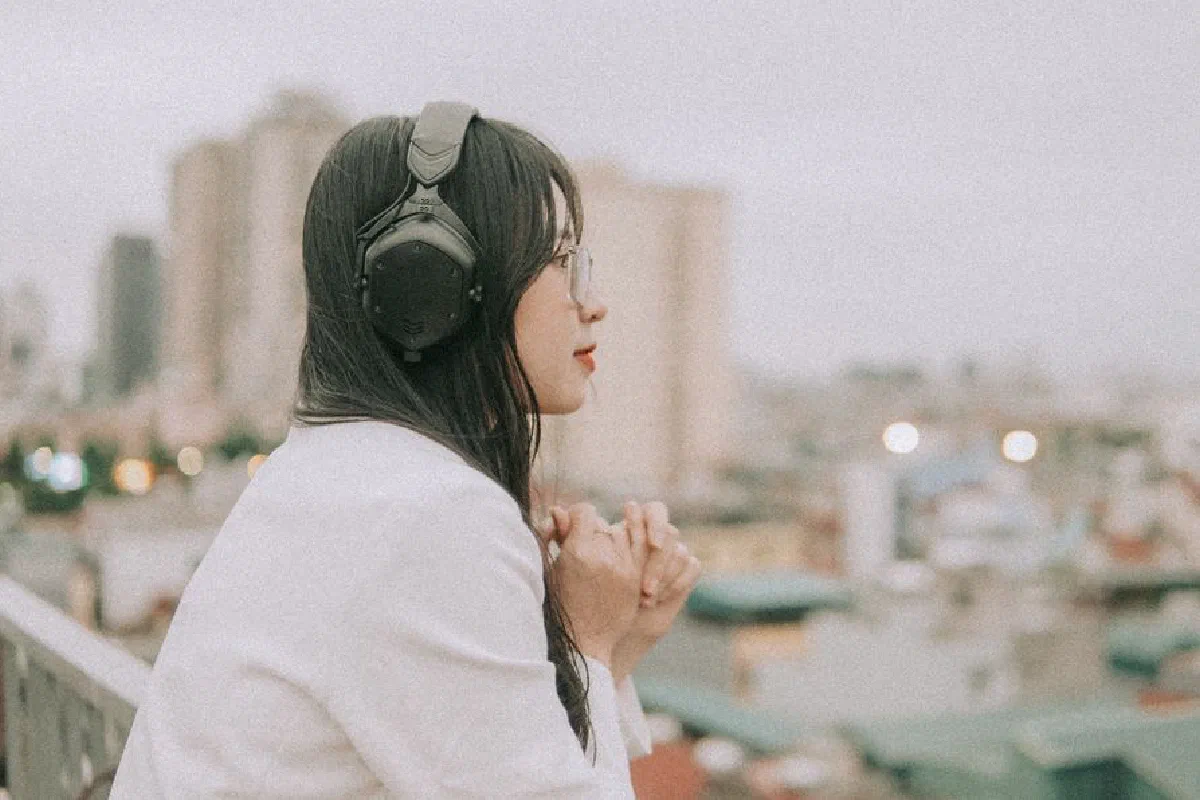 Side view of a woman in headphones and eyeglasses, enjoying a cityscape from a rooftop.
