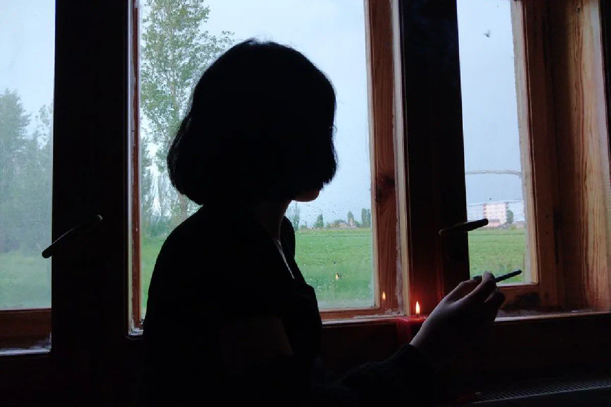 A silhouette of a person holding a cigarette by a window on a rainy day.