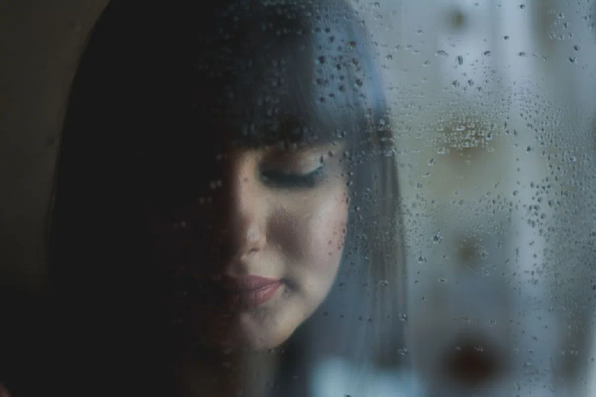A woman's thoughtful expression through a rain-soaked window creates a moody, introspective atmosphere.