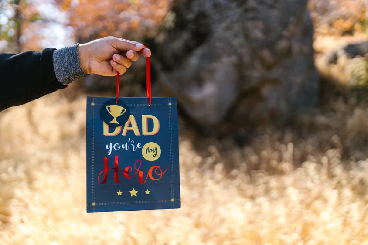 A hand holds a gift bag with 'Dad, you're my hero' text in a natural setting.