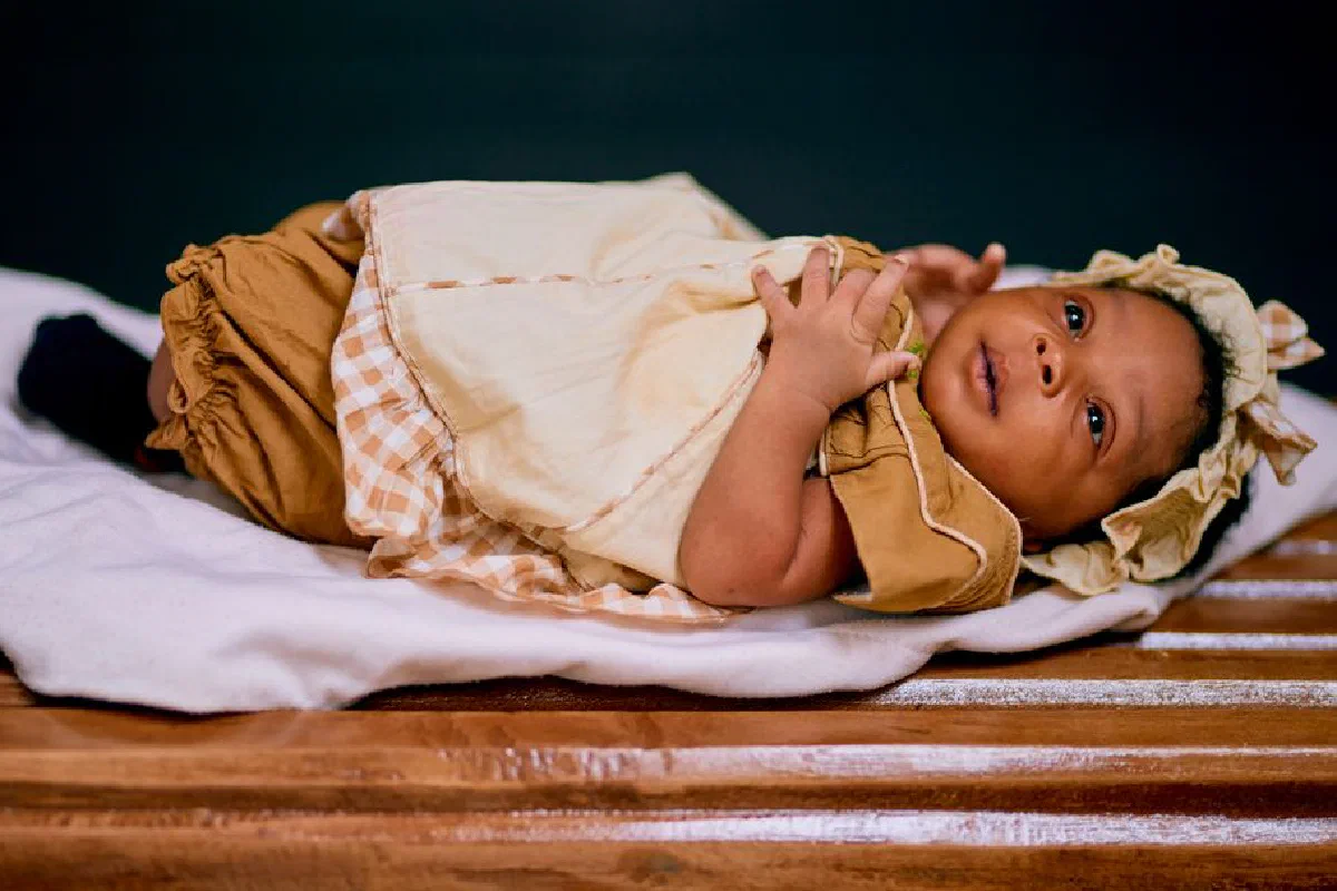 Adorable African baby in traditional beige outfit lying on a blanket indoors.