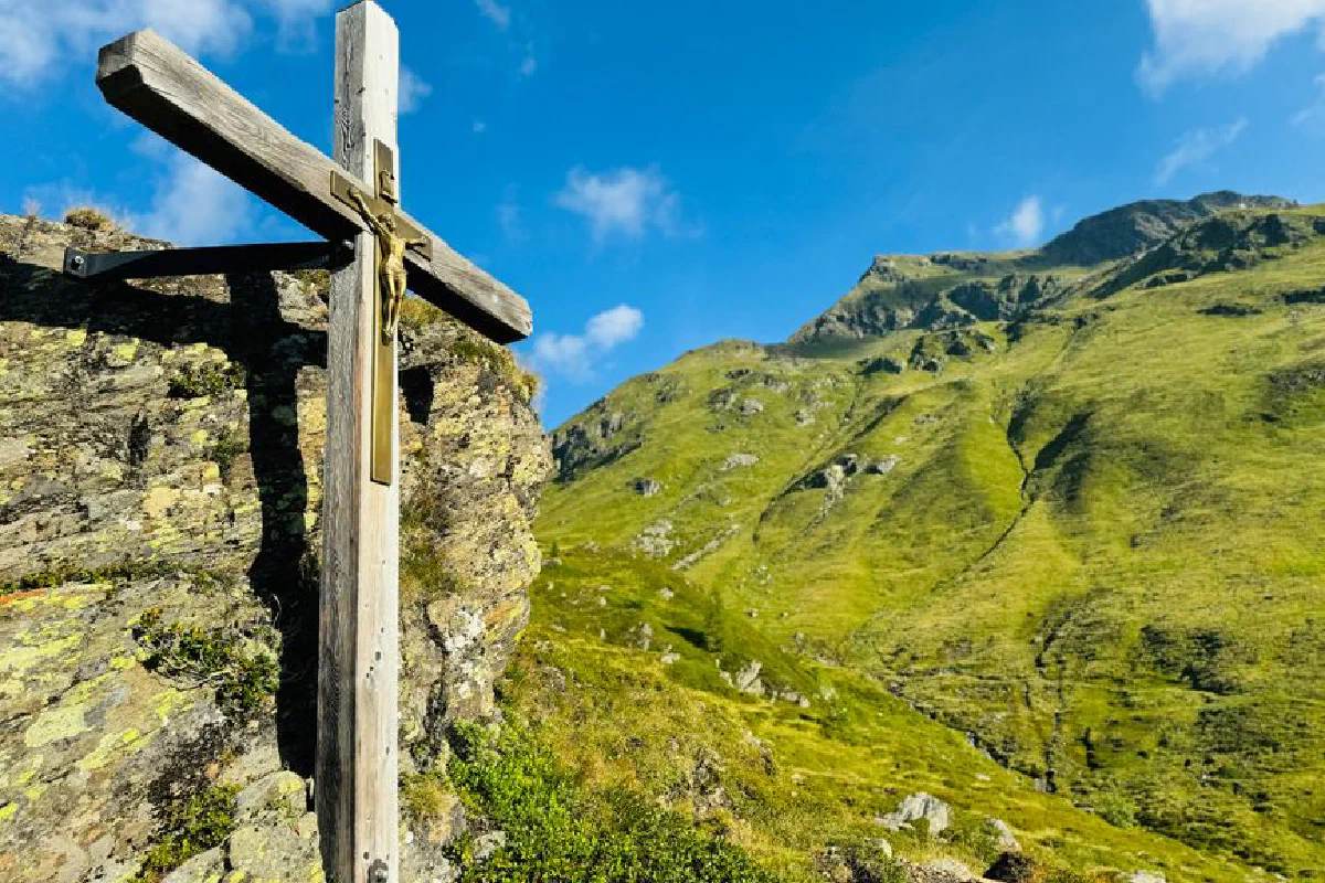 A wooden cross stands on a rocky hill with a mountain backdrop under a clear blue sky.