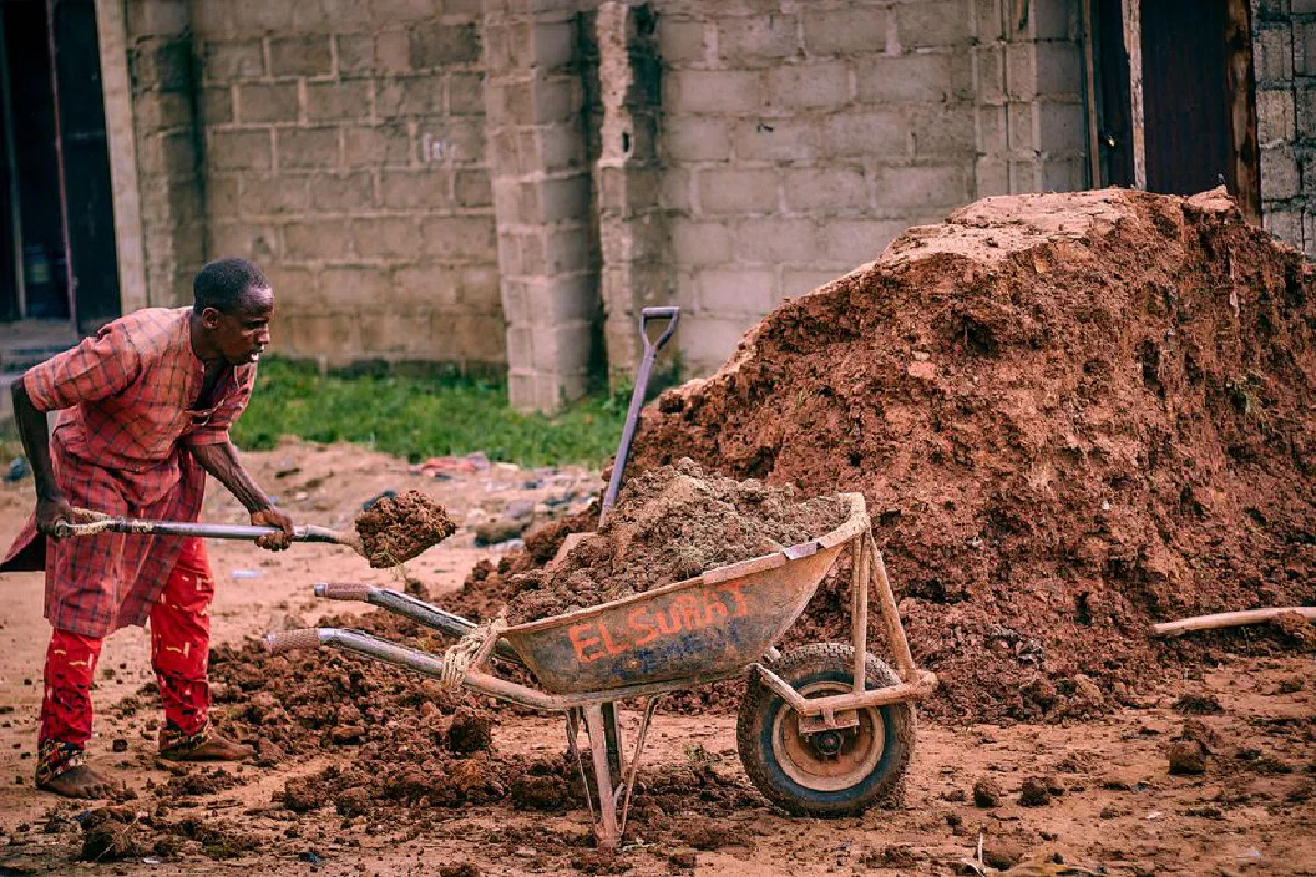 An African man laboring with dirt and a wheelbarrow in a construction area.
