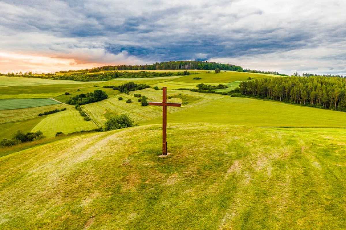 Aerial view of a lone wooden cross on a verdant hillside with dramatic sunset sky.