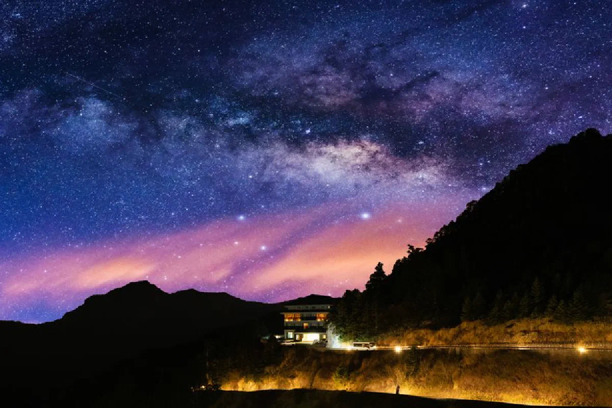 Stunning view of the Milky Way over a serene mountain landscape with a lit house at night.